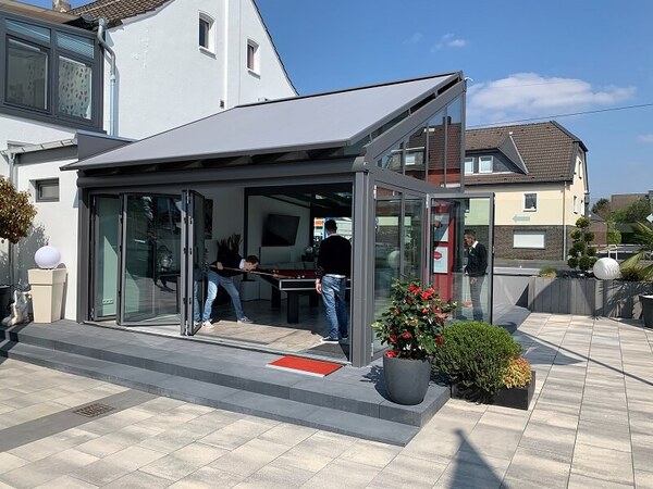 Moderner Wintergarten mit Glasdach, geöffneten Schiebetüren und Menschen beim Billardspiel.